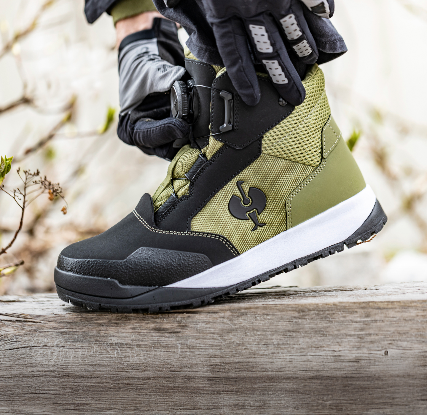 A gloved hand tightens a strap on an olive green and black hiking boot resting on a wooden surface, with an outdoor background.