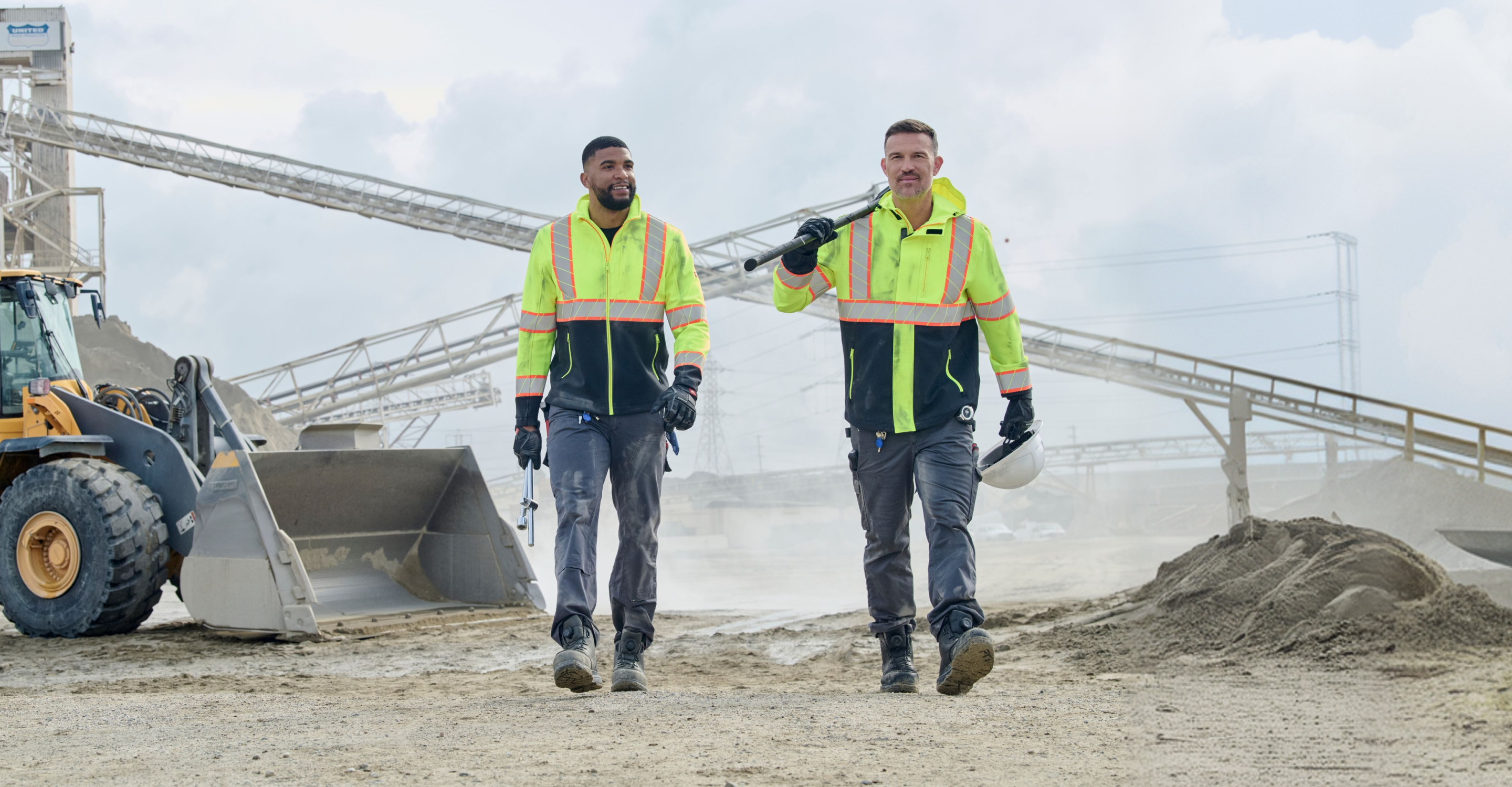 Two construction workers in high-visibility jackets walk on a sandy site, with industrial machinery and conveyor belts in the background on a cloudy day.