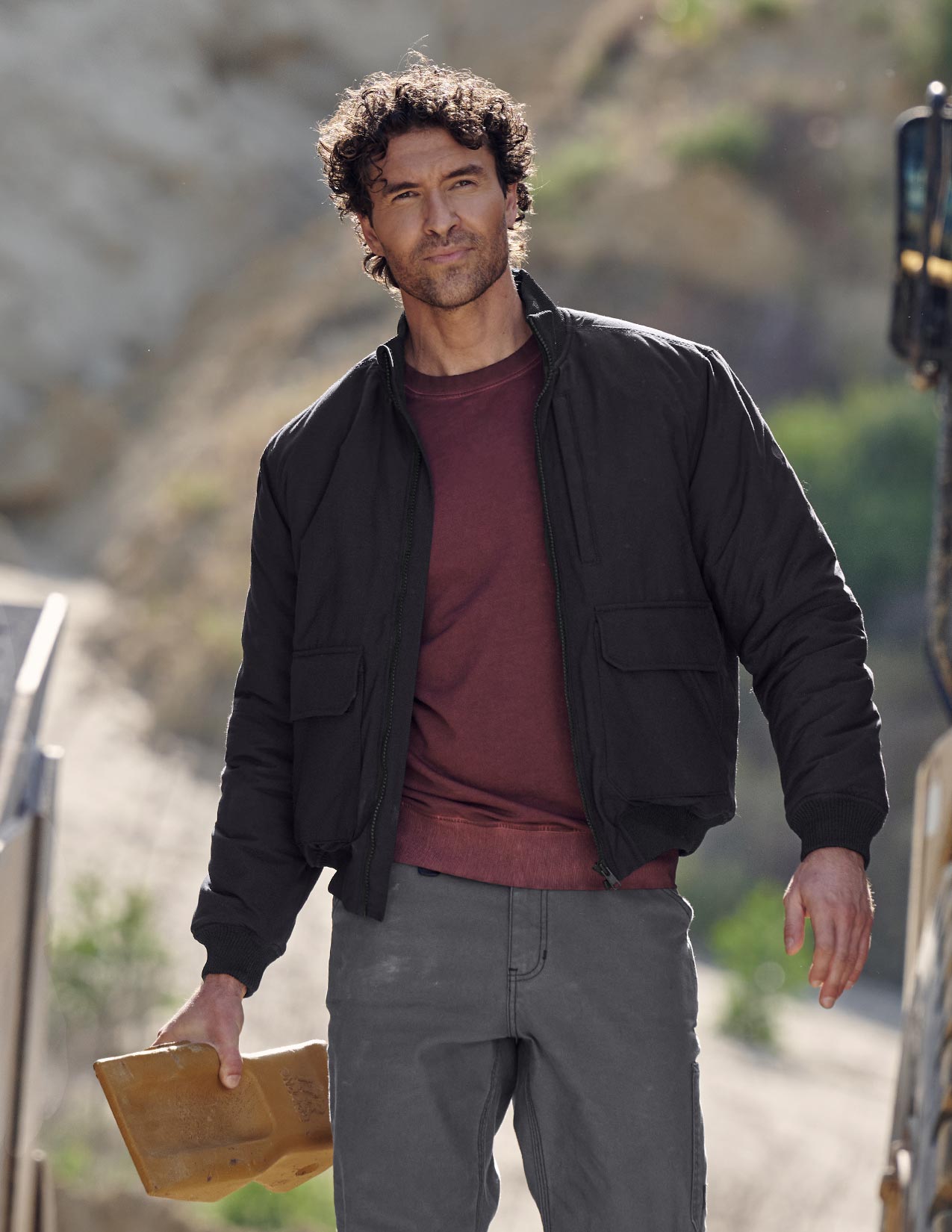 Man wearing a black jacket over a red shirt, holding a yellow object, with a blurred natural background