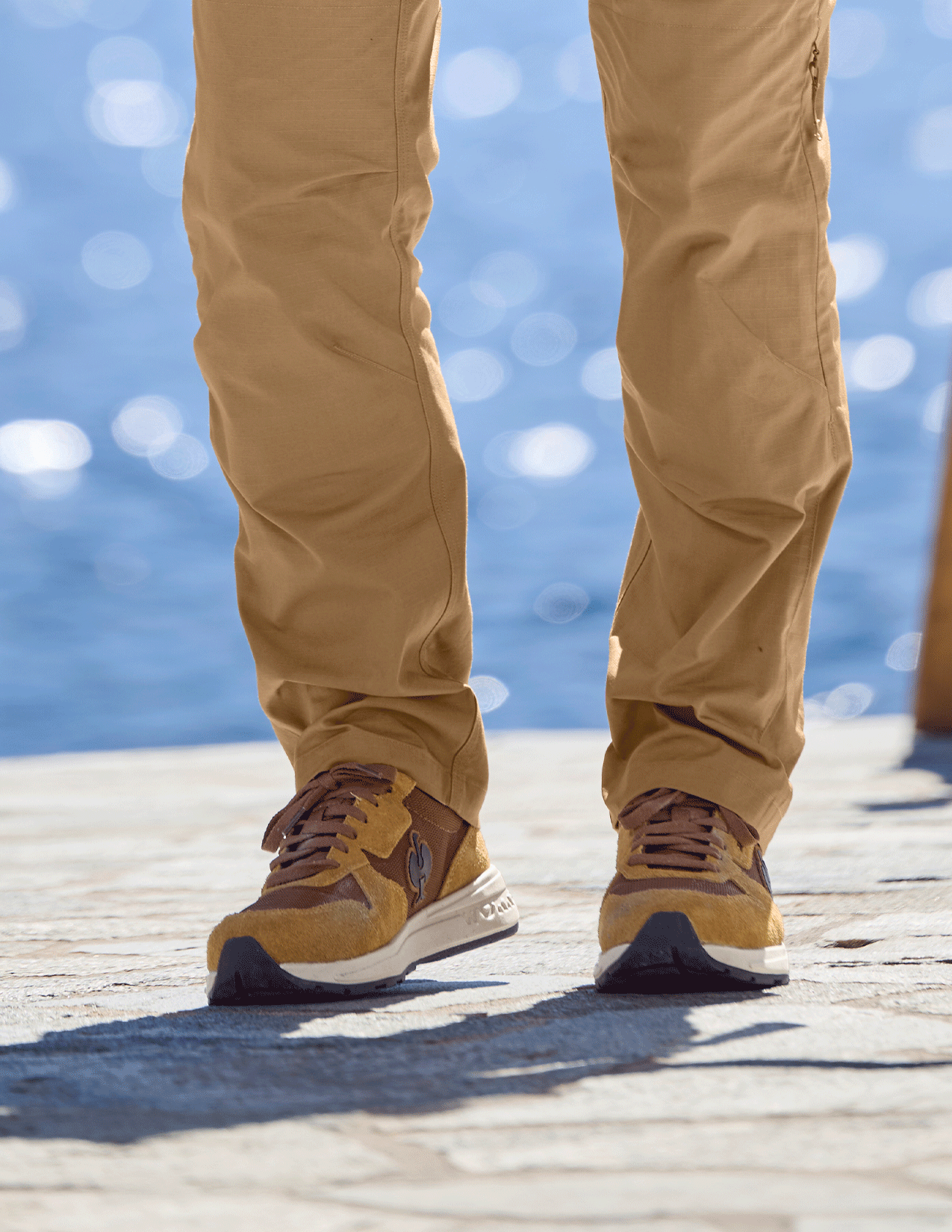 Person wearing desertbrown shoes and pants standing on a wooden dock with water in the background