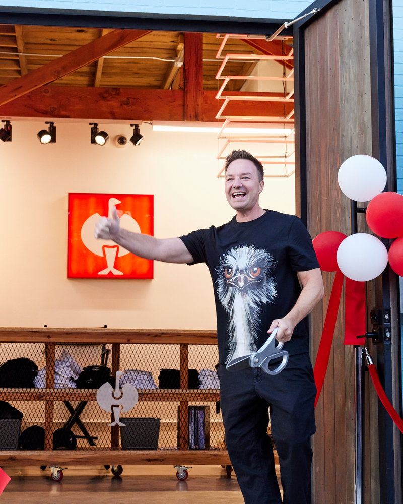 A smiling person holding large scissors stands at an open doorway with red and white balloons, appearing to cut a red ribbon at an event. The person wears a t-shirt featuring a large ostrich face design.