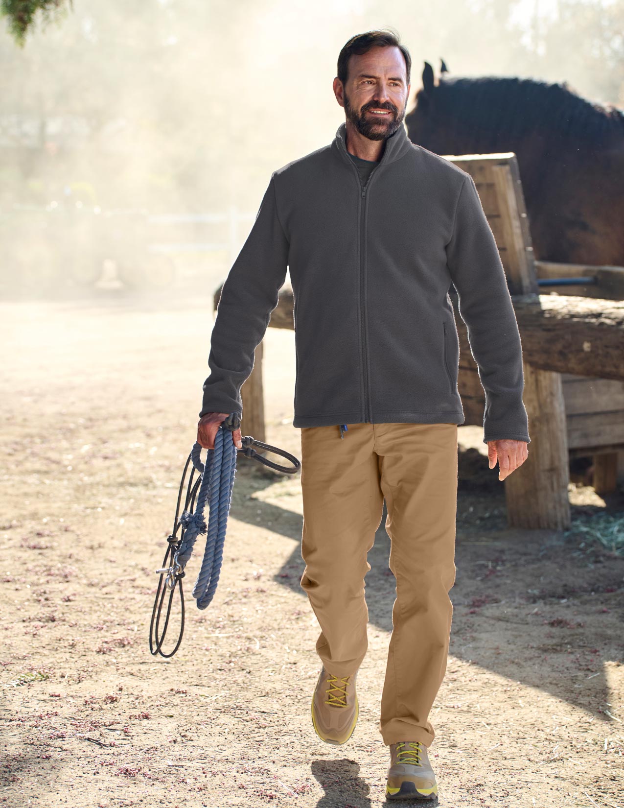 Man in a gray jacket and beige pants walking with a horse in the background