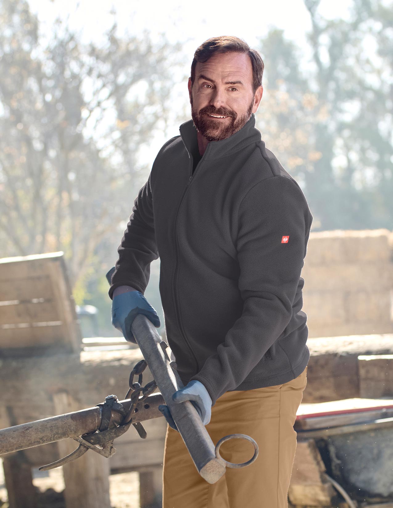 Man wearing a dark jacket with a logo, holding a tool outdoors.