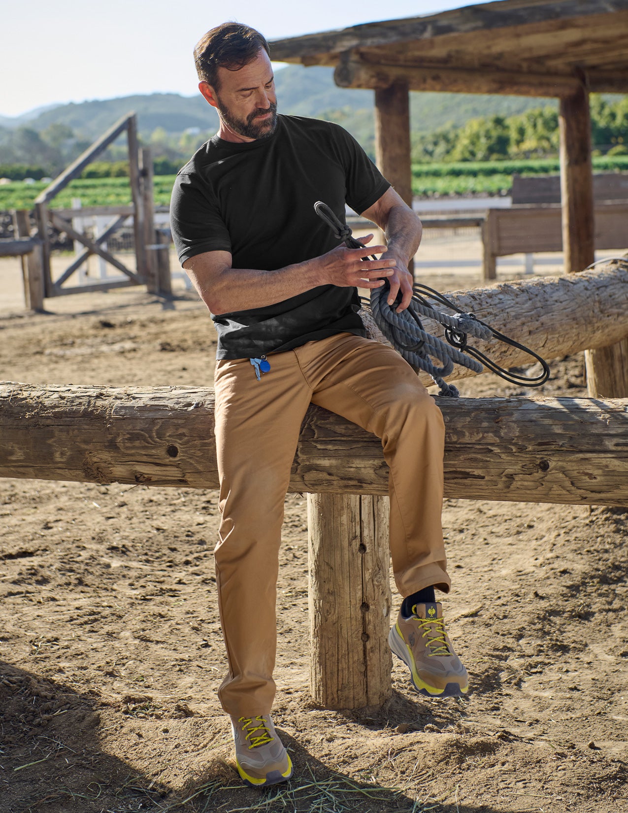 Man sitting on a wooden log outdoors with a rifle, wearing a black shirt and tan pants.