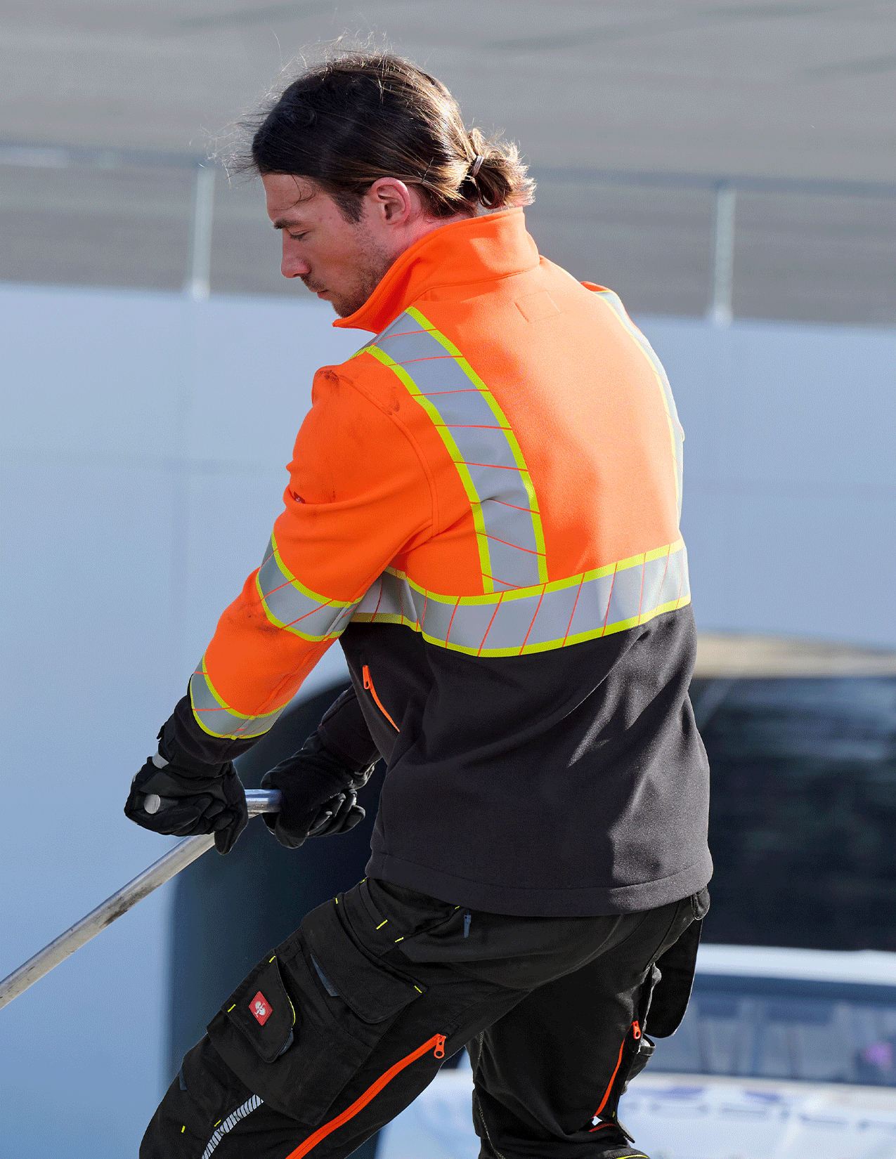 Person wearing an orange safety vest and black pants, holding a tool against a light blue wall.