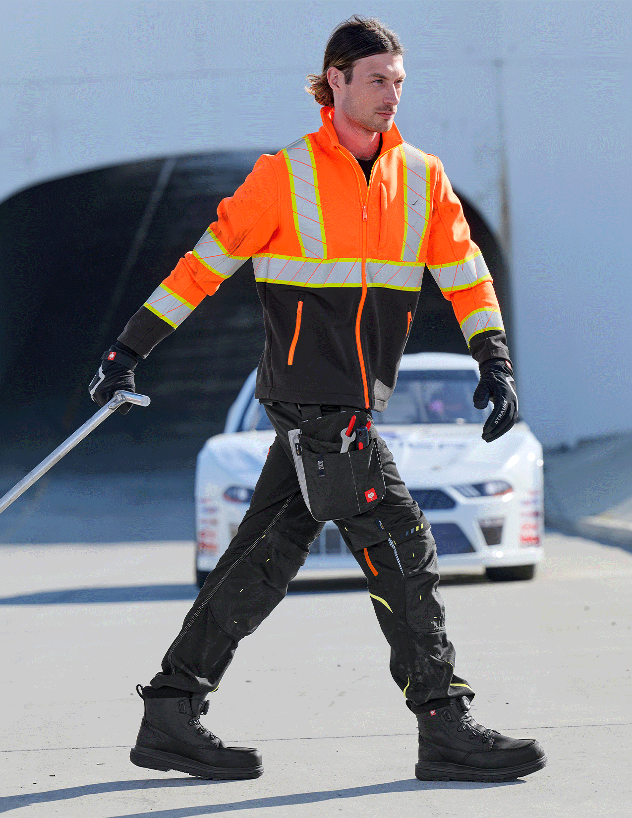 Person wearing an orange safety jacket with reflective stripes, black pants, and gloves, holding a tool on a road.