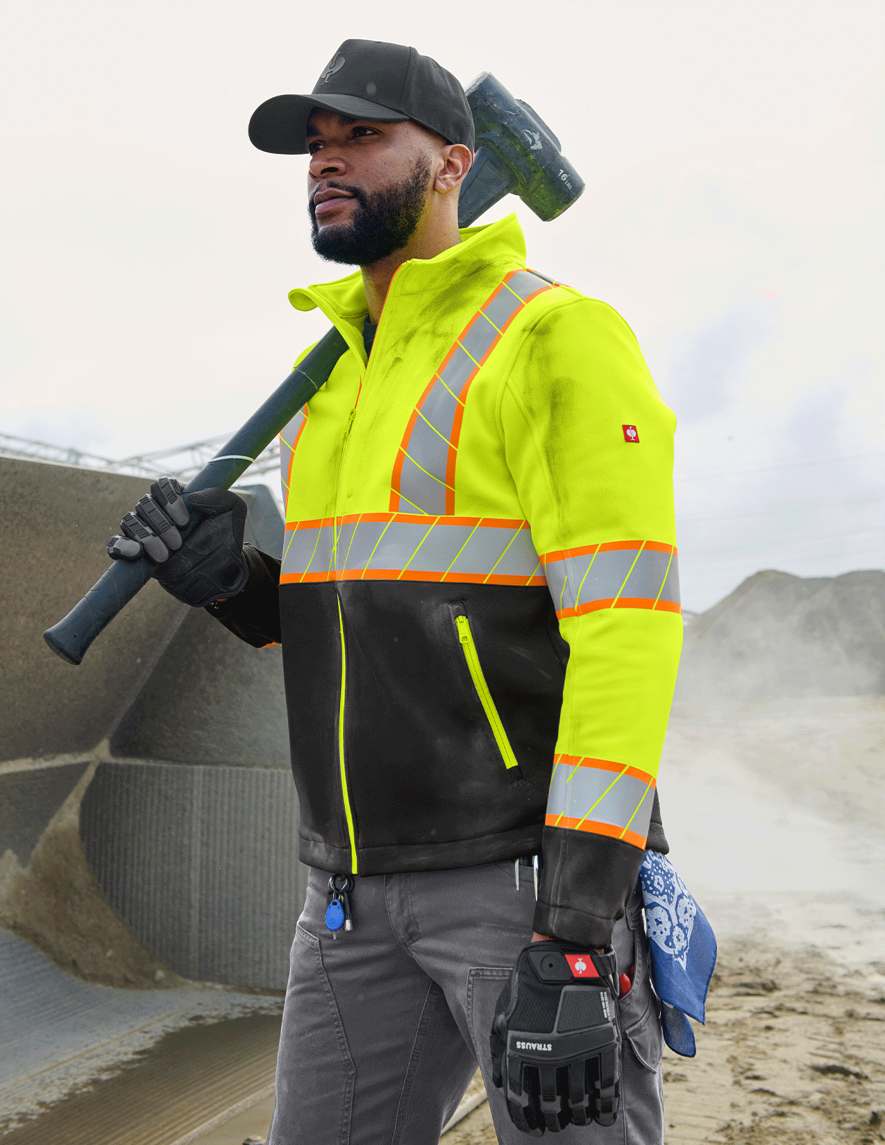 Person wearing a high-visibility safety jacket with reflective stripes, holding a tool against a construction site background.