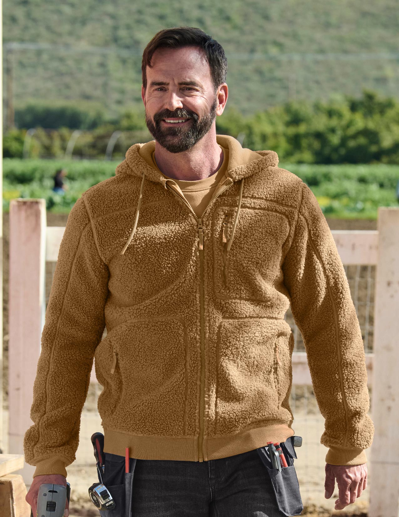Man wearing a brown fleece jacket with tools in his pockets, standing outdoors.
