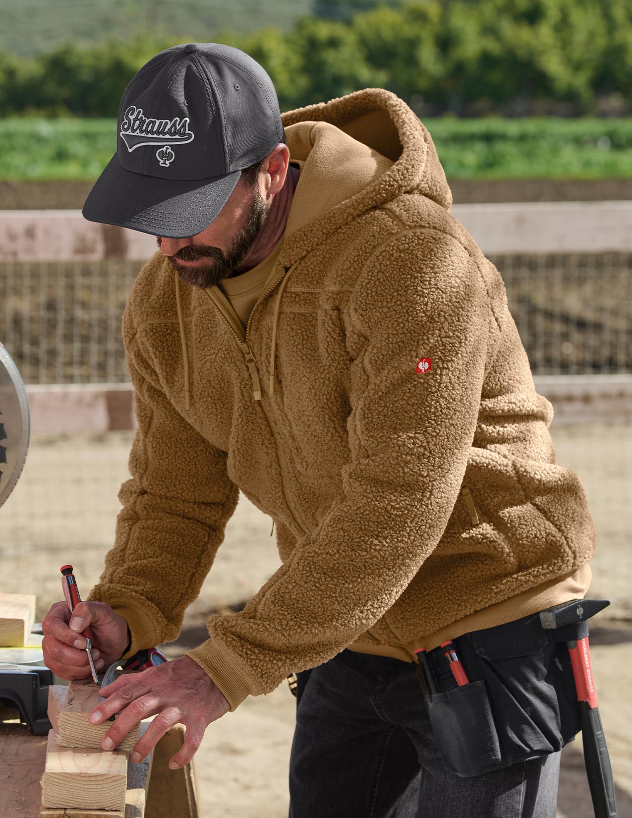 Man wearing a brown fleece jacket and grey cap working with wood outdoors.
