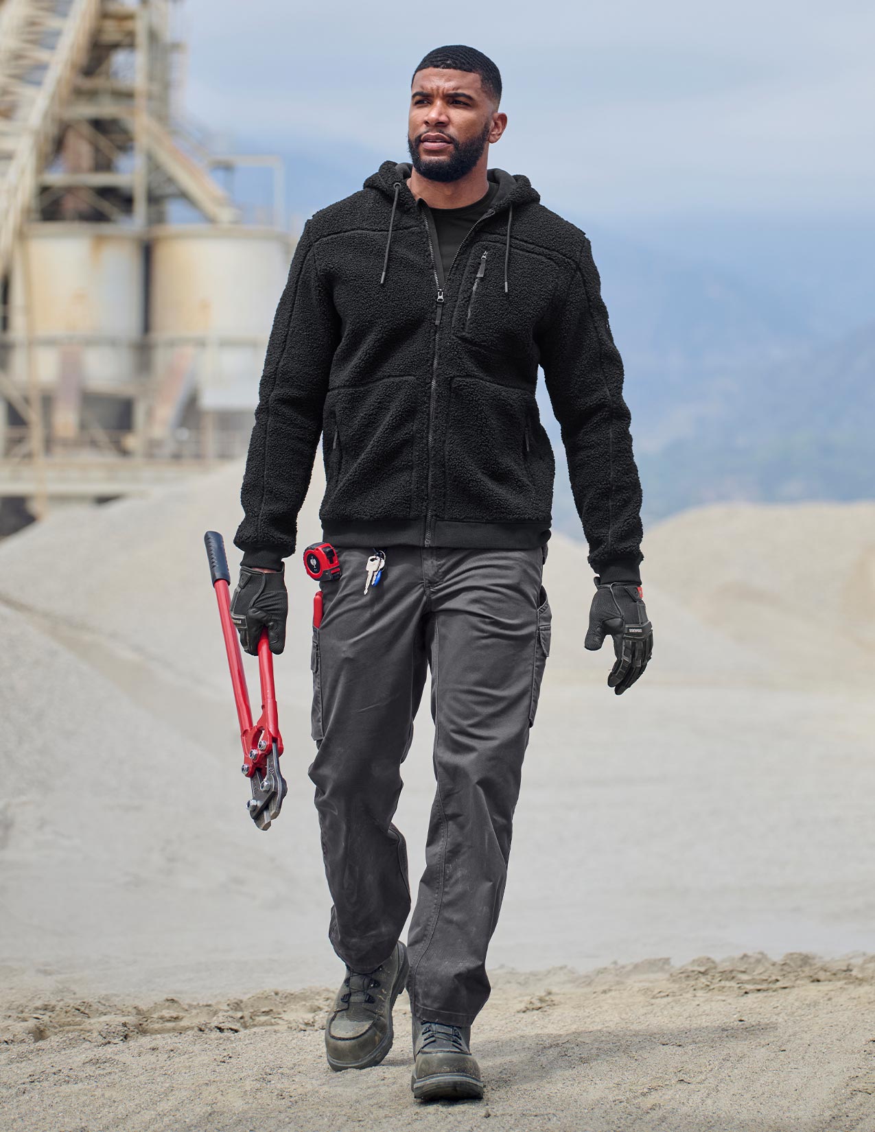 Person wearing a black jacket and gray pants holding tools on a construction site.