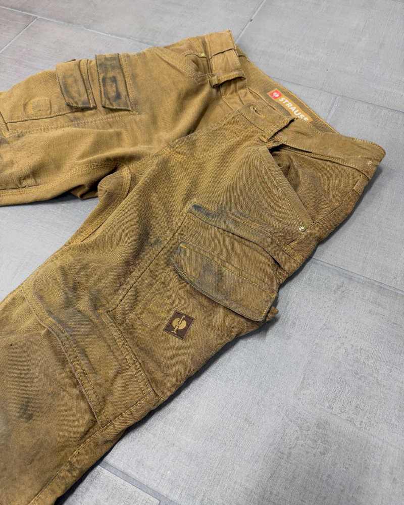 Brown work pants with multiple pockets and tool loops, showing visible black stains and signs of wear, are laid flat on a gray tiled floor.