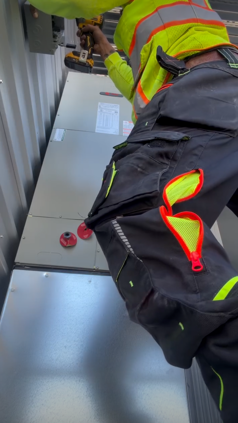 A worker in a neon safety vest and work pants uses a power drill on metal equipment mounted to a wall, possibly performing installation or maintenance. Two red caps are visible on the equipment beside the worker.