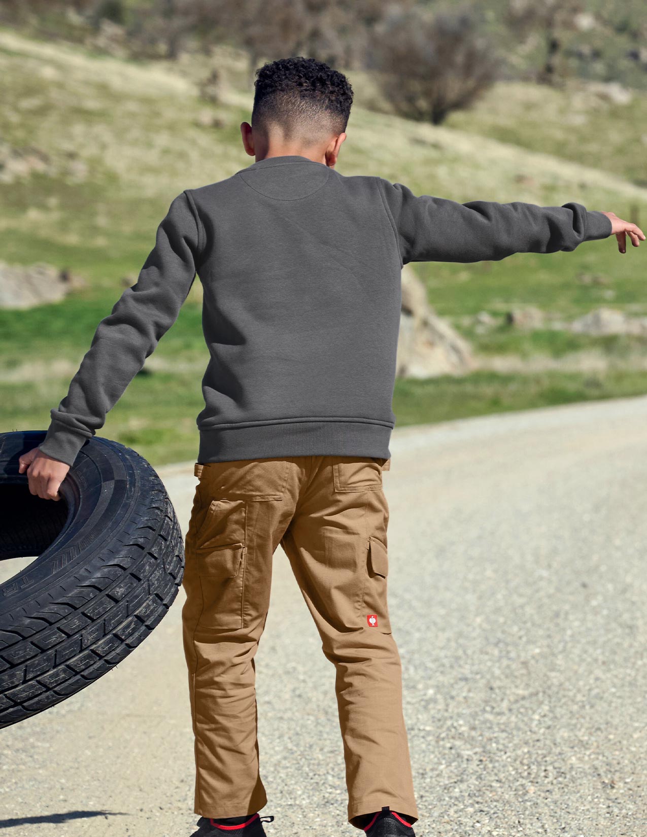 Child wearing a gray sweatshirt and brown pants holding a tire on a road with a natural landscape in the background.