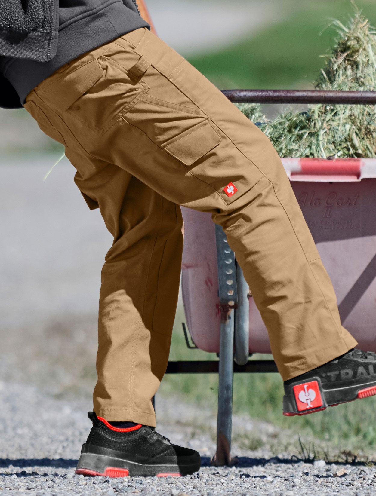 Child wearing brown pants and black boots with red accents, standing near a wheelbarrow.