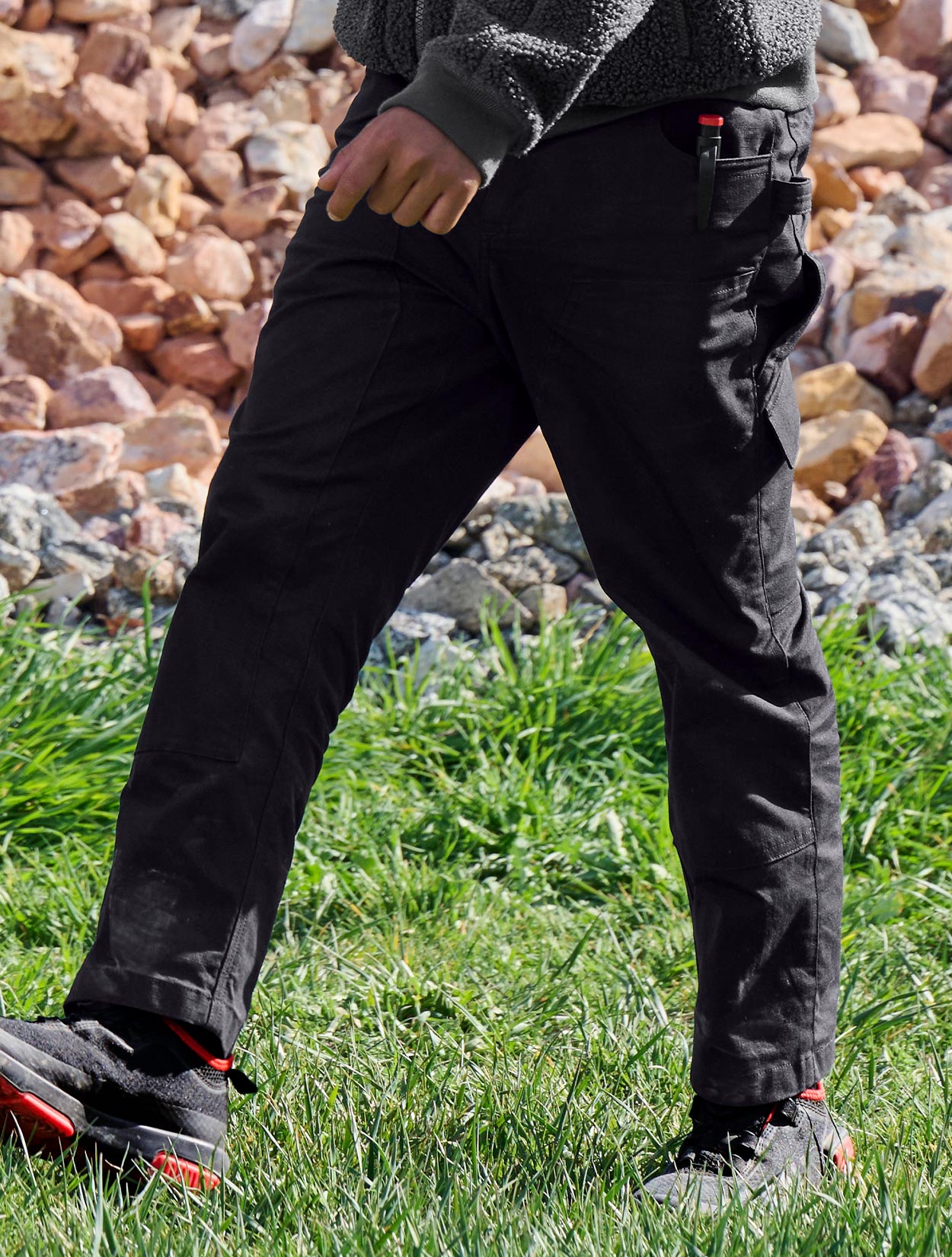 Child wearing black pants and shoes standing on grass with a rocky background