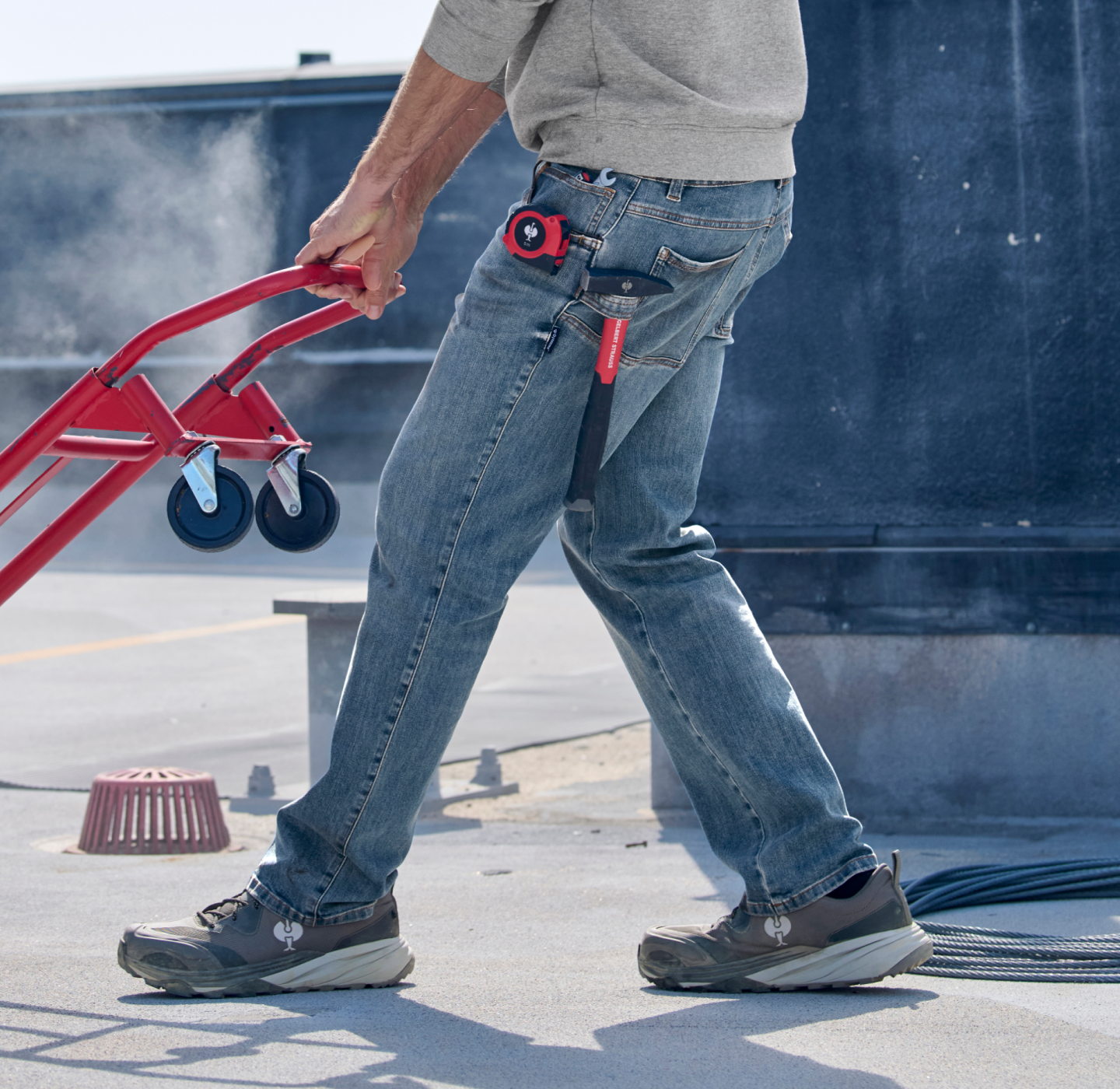 A person in blue jeans and gray shoes pushes a red hand truck outdoors. A utility knife and tape measure are clipped to their pocket. Steam or dust is visible in the background.
