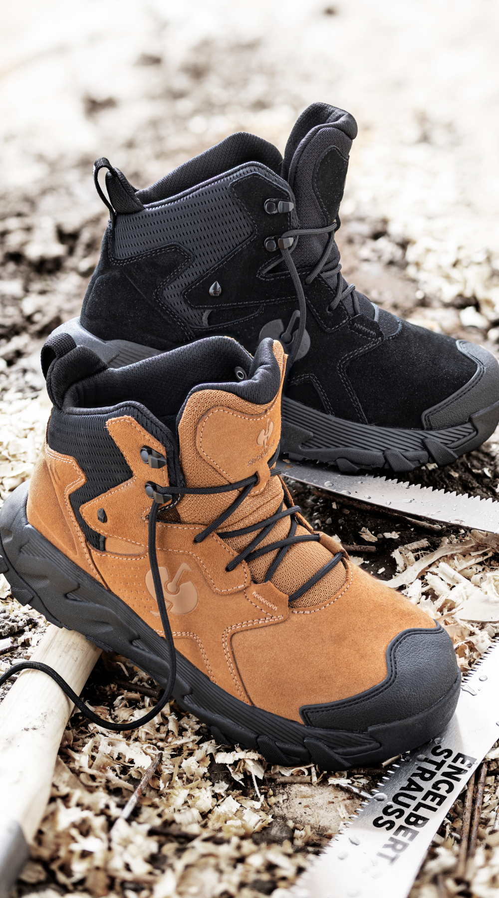 Two rugged hiking boots, one tan and one black, sit side by side on wood shavings and dirt, next to a partially visible saw with a wooden handle and STAINLESS etched on the metal blade.