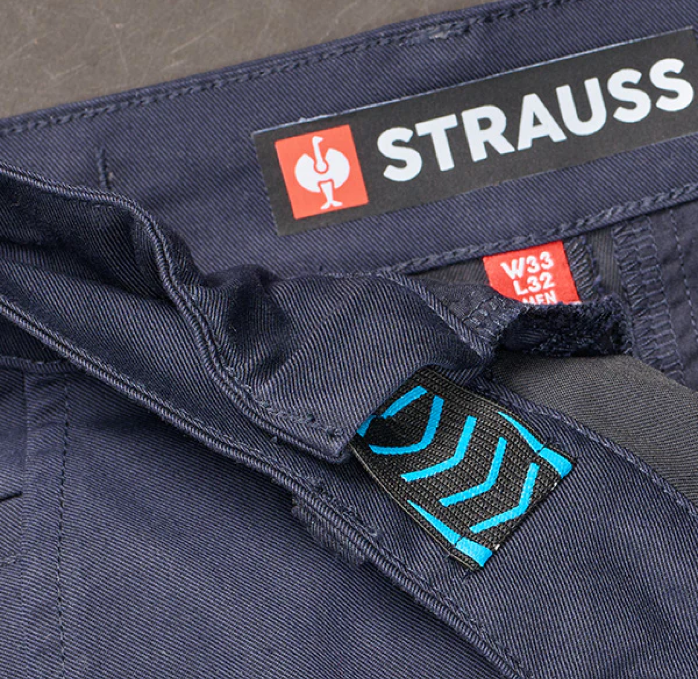 Close-up of a navy-blue garments waistband showing a STRAUSS brand label, a red size tag reading W33 L32, and a blue-accented loop with stitched chevrons.