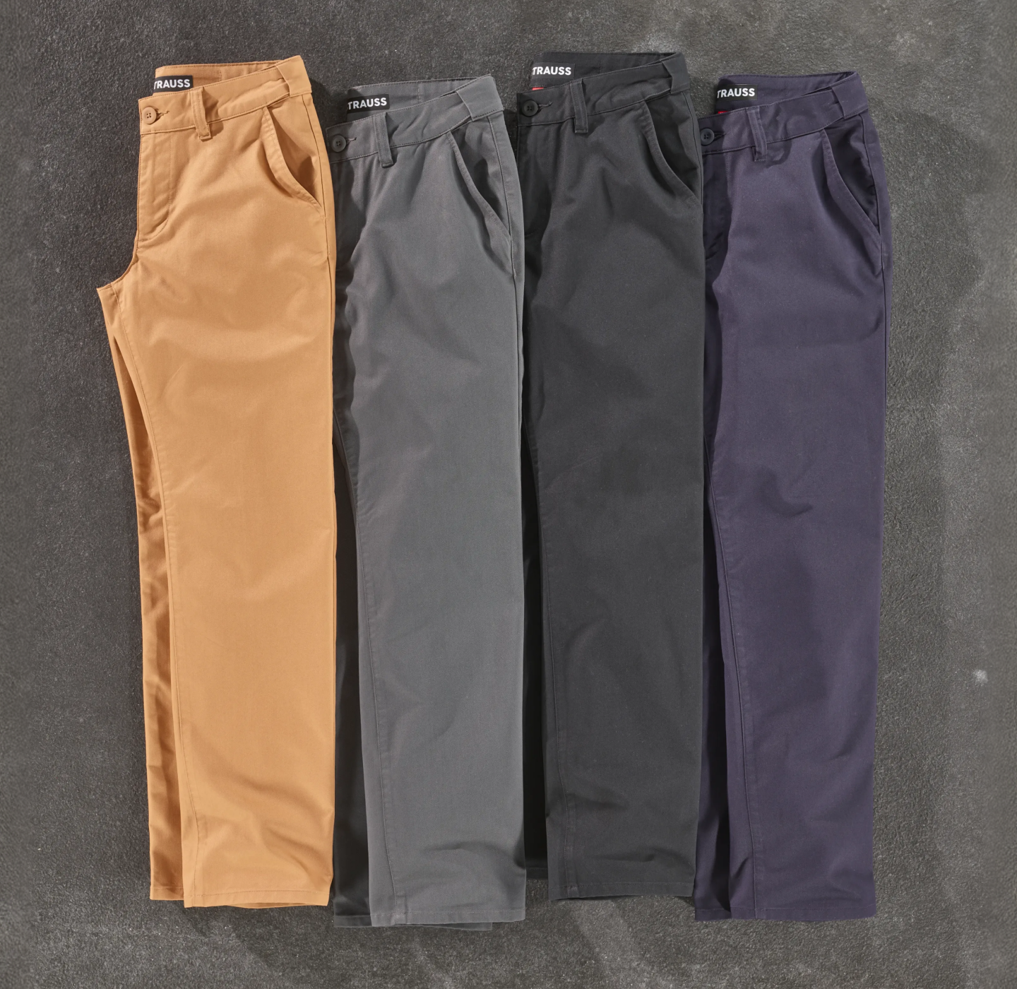 Four pairs of neatly folded pants in tan, gray, black, and navy colors are laid side by side on a textured gray surface.
