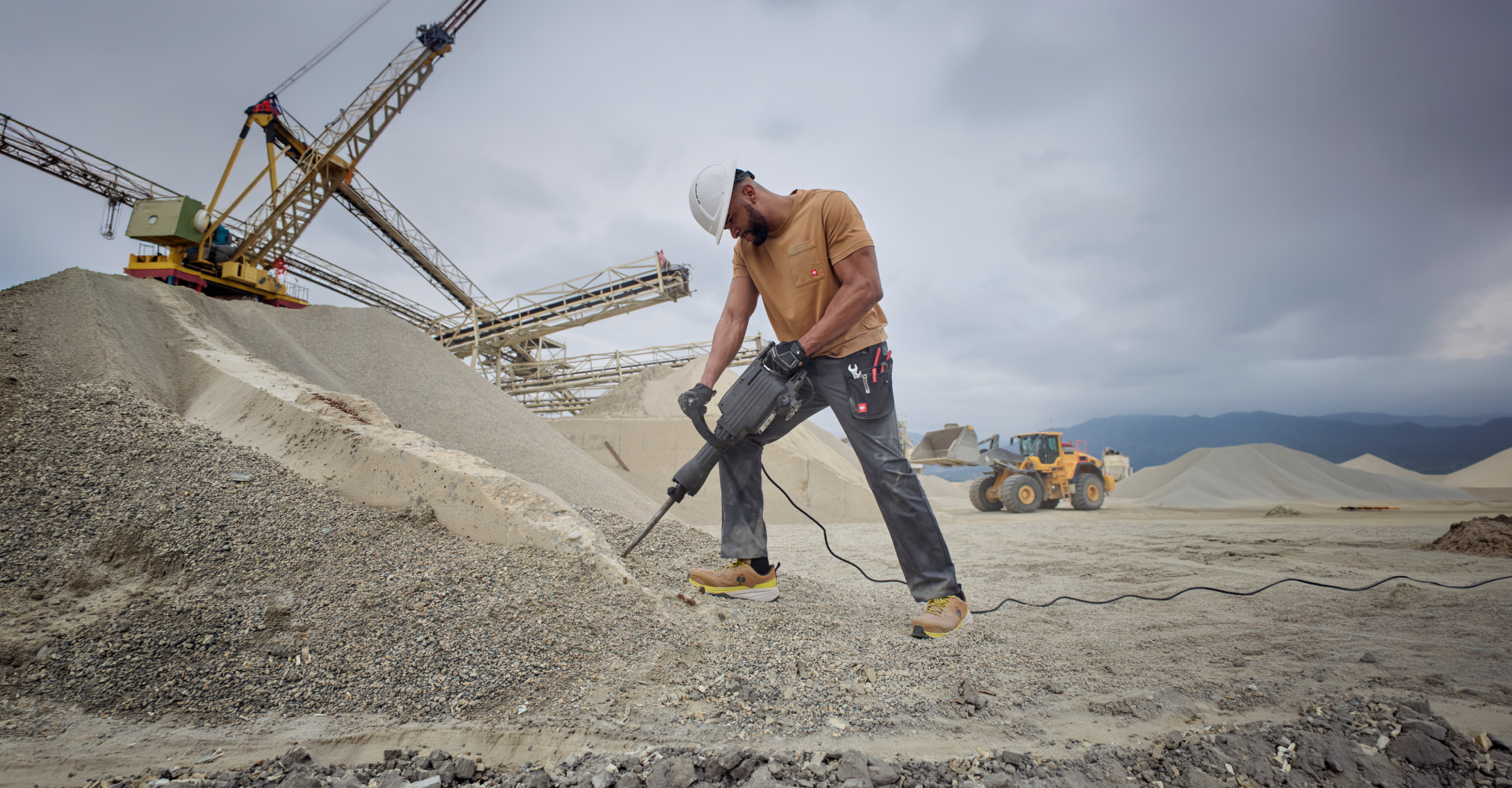 A construction worker in a hard hat uses a jackhammer on gravel at a quarry, with mounds of sand, heavy machinery, and cloudy sky in the background.