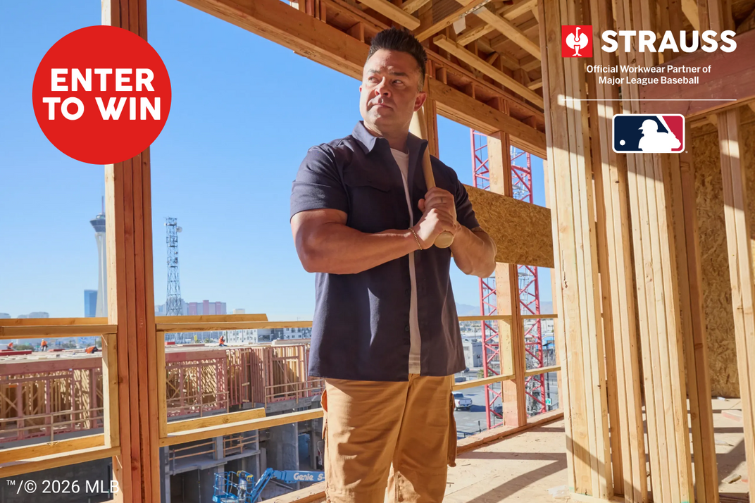 A man stands on an unfinished wooden construction site wearing a dark navy short-sleeve button-up shirt and beige work pants. Prominent overlays include a red 'ENTER TO WIN' badge, Strauss Official Worker Partner of MLB Baseball branding, and MLB copyright text.