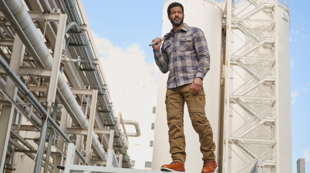 A man wearing a plaid button-up shirt, brown cargo work pants e.s.vintage, and orange sneakers stands on a platform in front of large white industrial tanks and pipes under a blue sky.