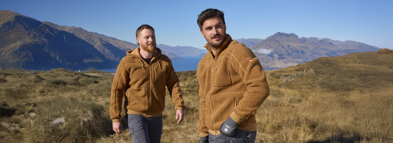 Two men wearing tan sherpa fleece jackets are standing in a mountain landscape with a lake and grasslands.