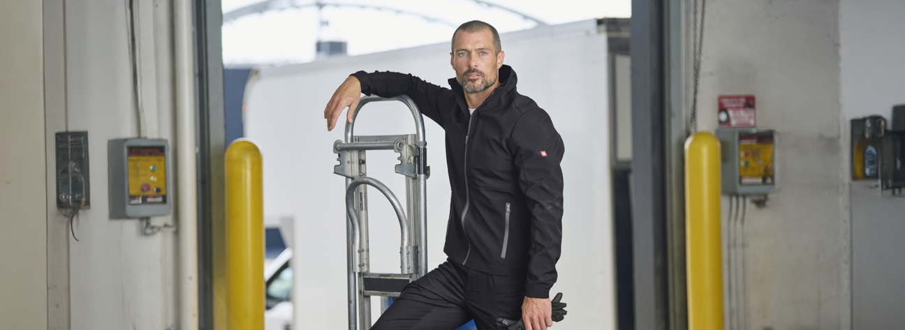 A man wearing a black softshell jacket with hood and black cargo pants is leaning on a folding ladder at an industrial garage with yellow safety bollards.