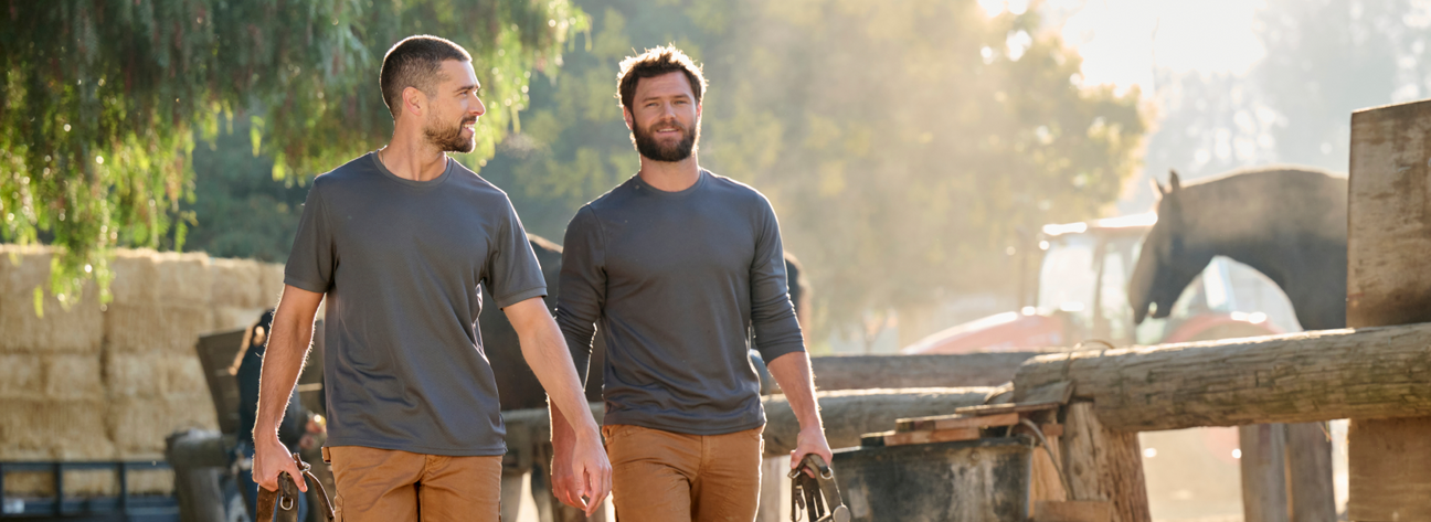 Two men wearing gray long-sleeve work shirts and tan pants are walking at an outdoor farm with trees and equipment.