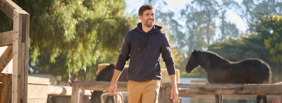 A man wearing a navy blue hoodie and tan cargo pants is walking at a ranch with wooden fencing and a horse in the background.