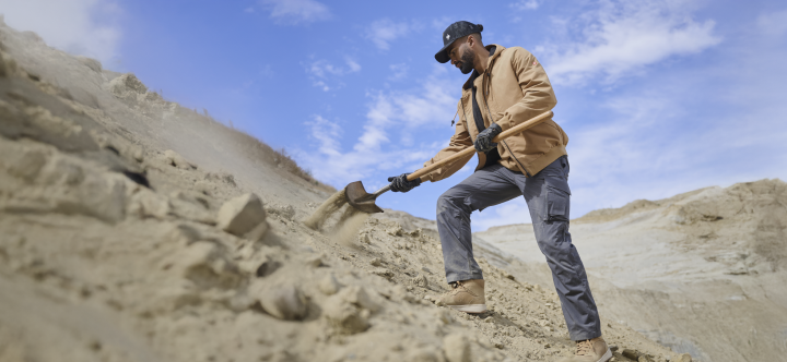 A man wearing a tan work jacket and gray work pants uses a sledgehammer on sandy terrain under a clear blue sky.