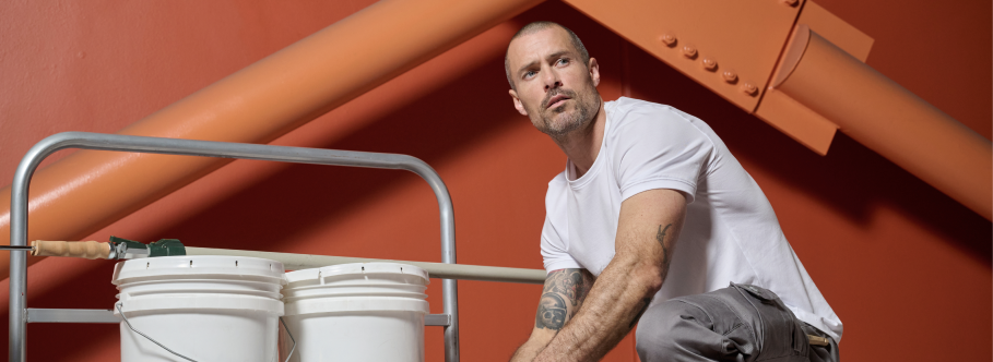 A male construction worker wearing a white t-shirt and gray work pants sits on an orange industrial structure.