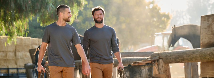 Two men wearing gray long-sleeve work shirts and tan pants are walking at an outdoor farm with trees and equipment.