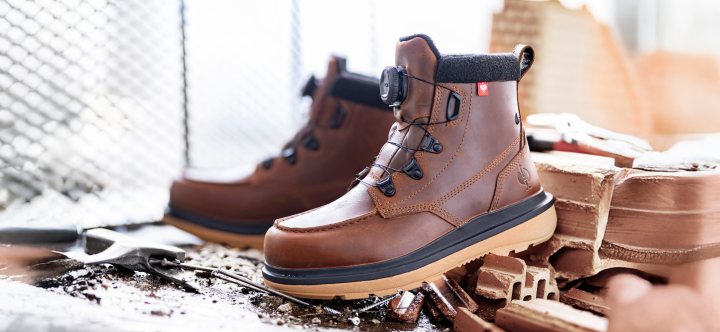 Brown leather safety toe work boots with BOA closure systems displayed on a wooden surface surrounded by work materials and supplies.