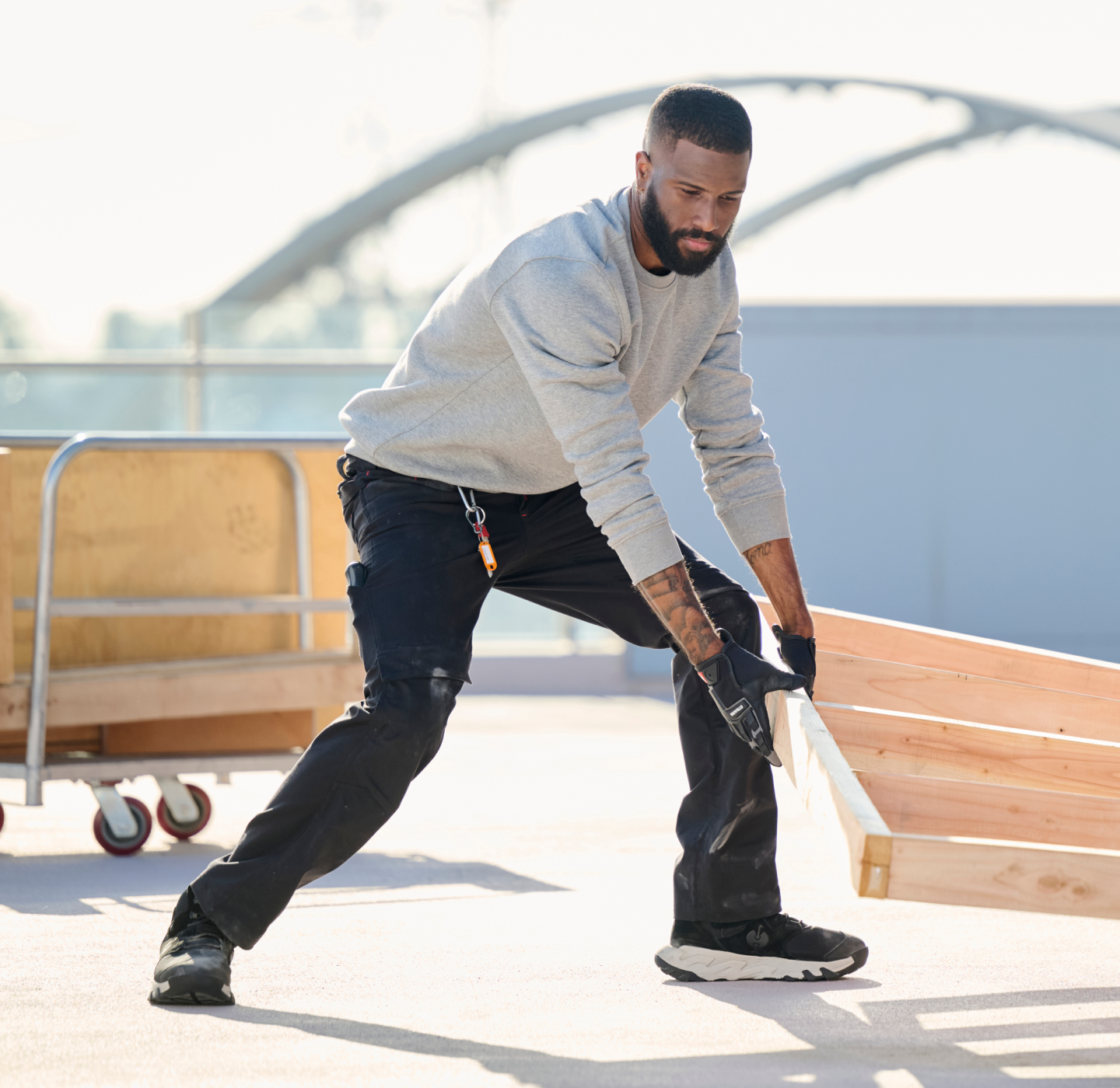 A man wearing gloves and casual work clothes lifts a wooden pallet on a sunny outdoor surface, with a metal cart and industrial background visible behind him.