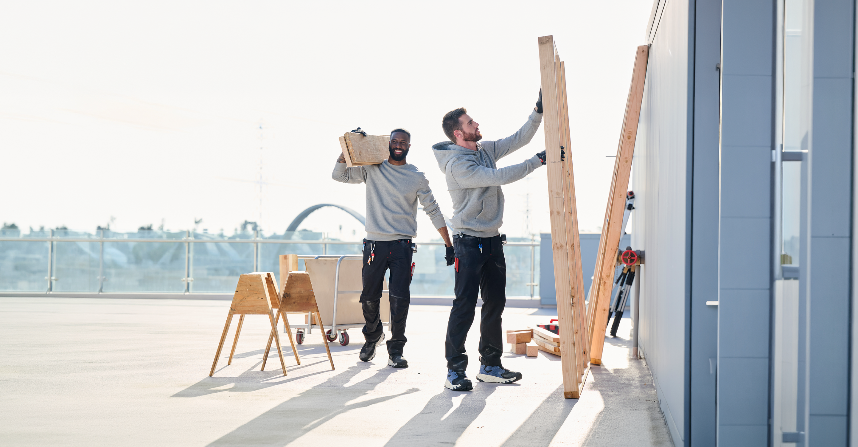 Two men wearing gray sweatshirts and work gloves are working on a rooftop. One lifts wooden beams against a wall, while the other carries a wooden box. Tools and a workbench are in the background.