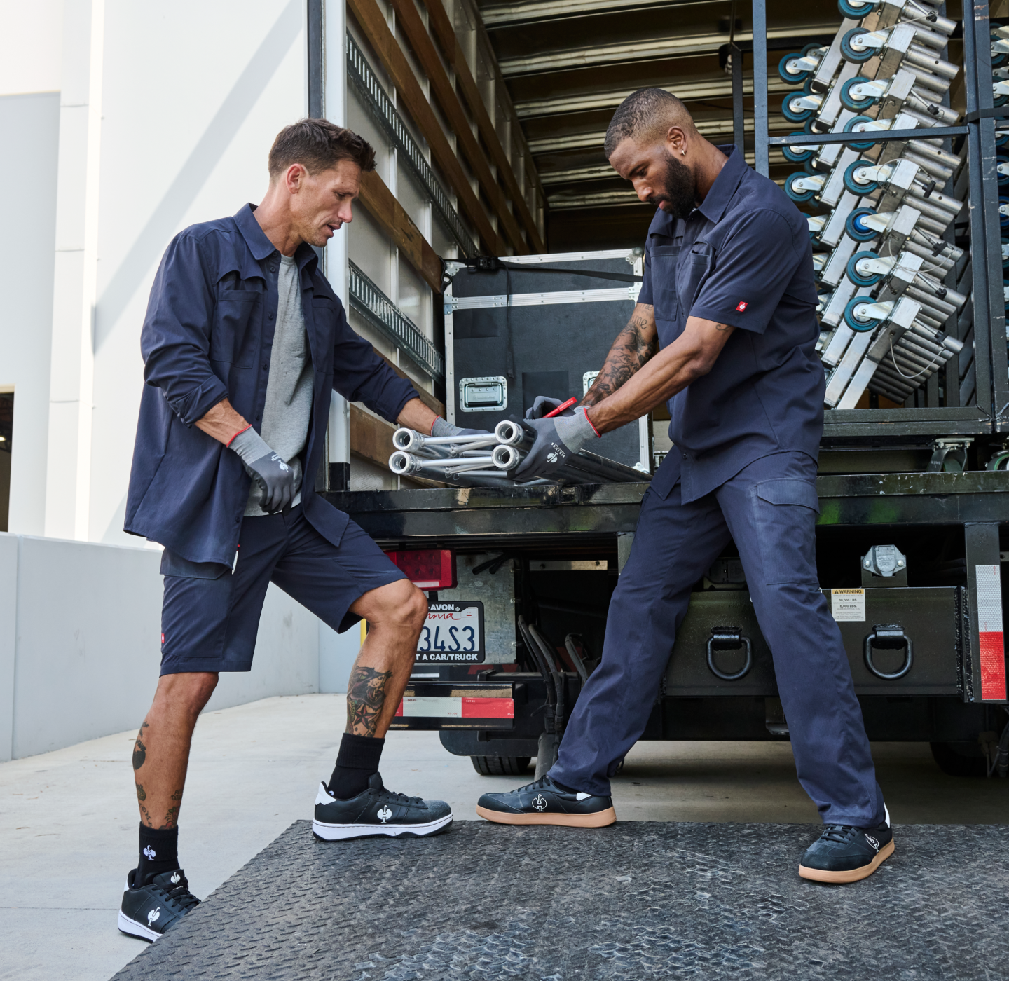 Two workers wearing dark uniforms unload metal pipes from a truck loading bay. One stands on the ground, the other on the truck, both working together and wearing gloves. Industrial equipment is visible in the truck.