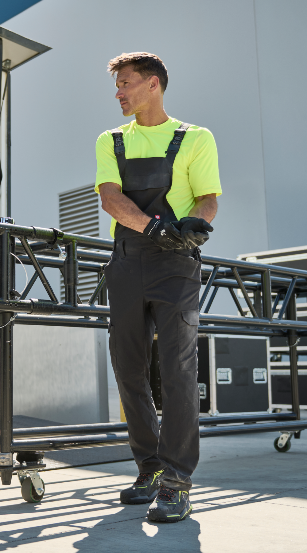 A man in a neon yellow shirt and black overalls stands outdoors next to metal equipment, wearing work gloves and looking sideways on a sunny day.
