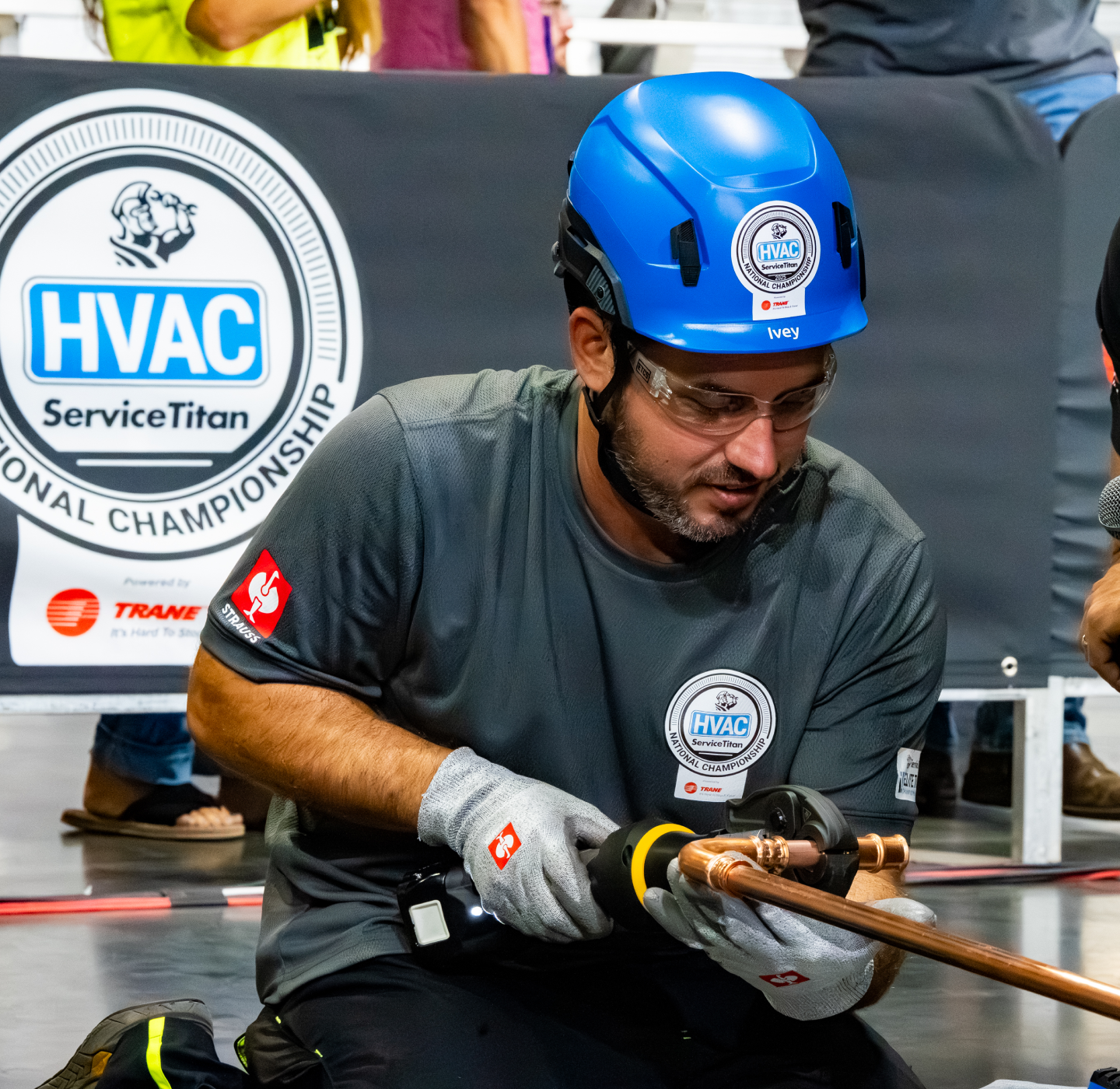 A man wearing safety gear works with copper pipes at an HVAC competition. He uses a tool while kneeling, and the event logo is visible in the background.