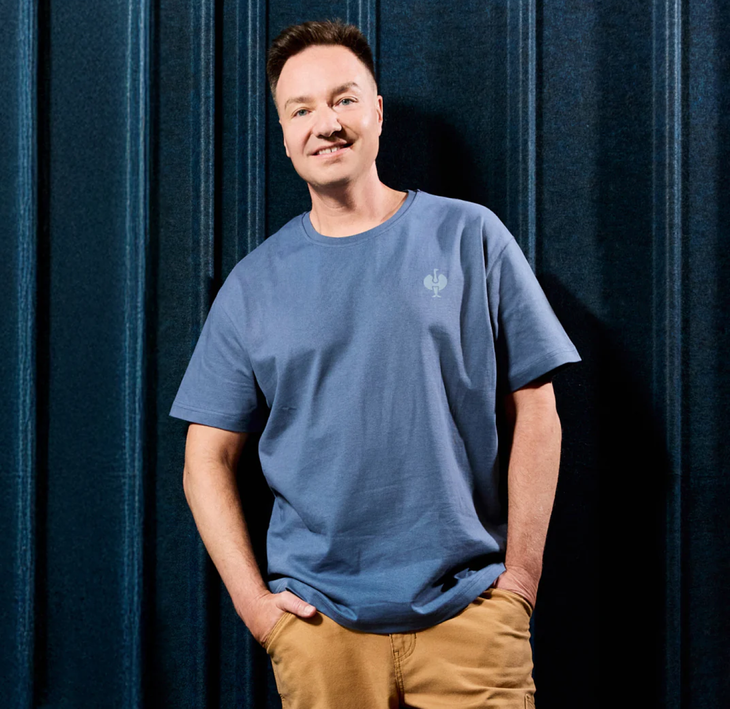 A smiling man stands in front of a dark blue textured wall, wearing a light blue t-shirt and tan pants with his hands in his pockets.