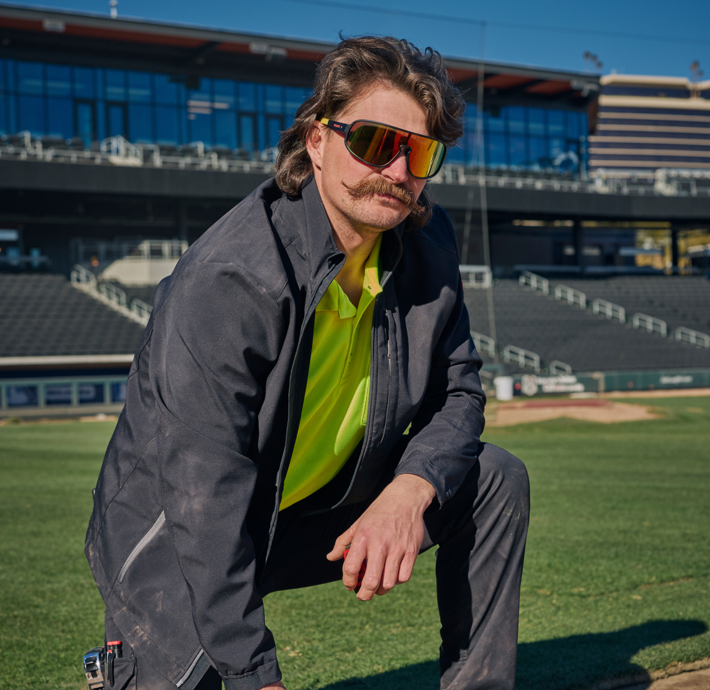 A man with a mustache wearing reflective sunglasses, a neon yellow shirt, and a dark jacket kneels on a baseball field, with empty stadium seats visible in the background.