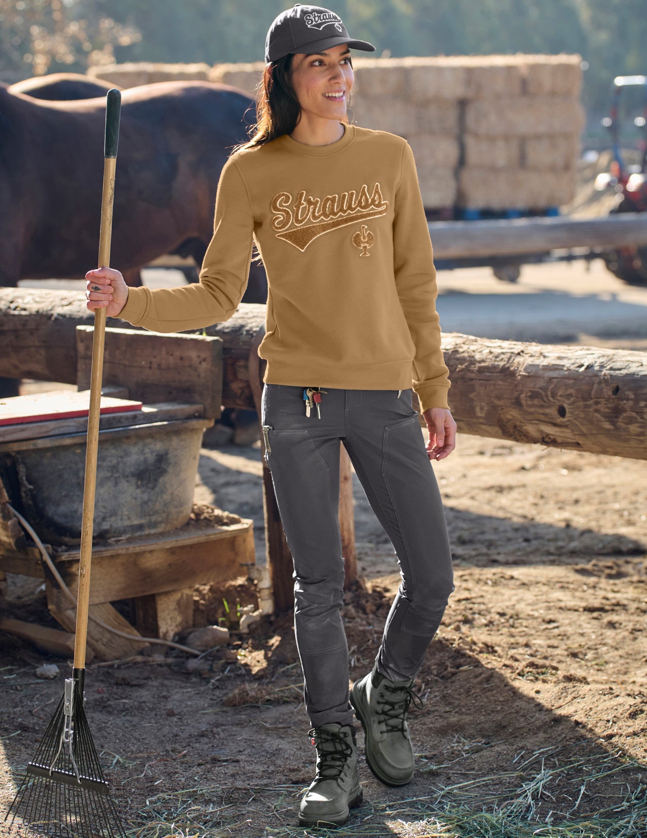 Woman in a yellow 'Strauss' sweatshirt on a farm with a cow and hay bales in the background.