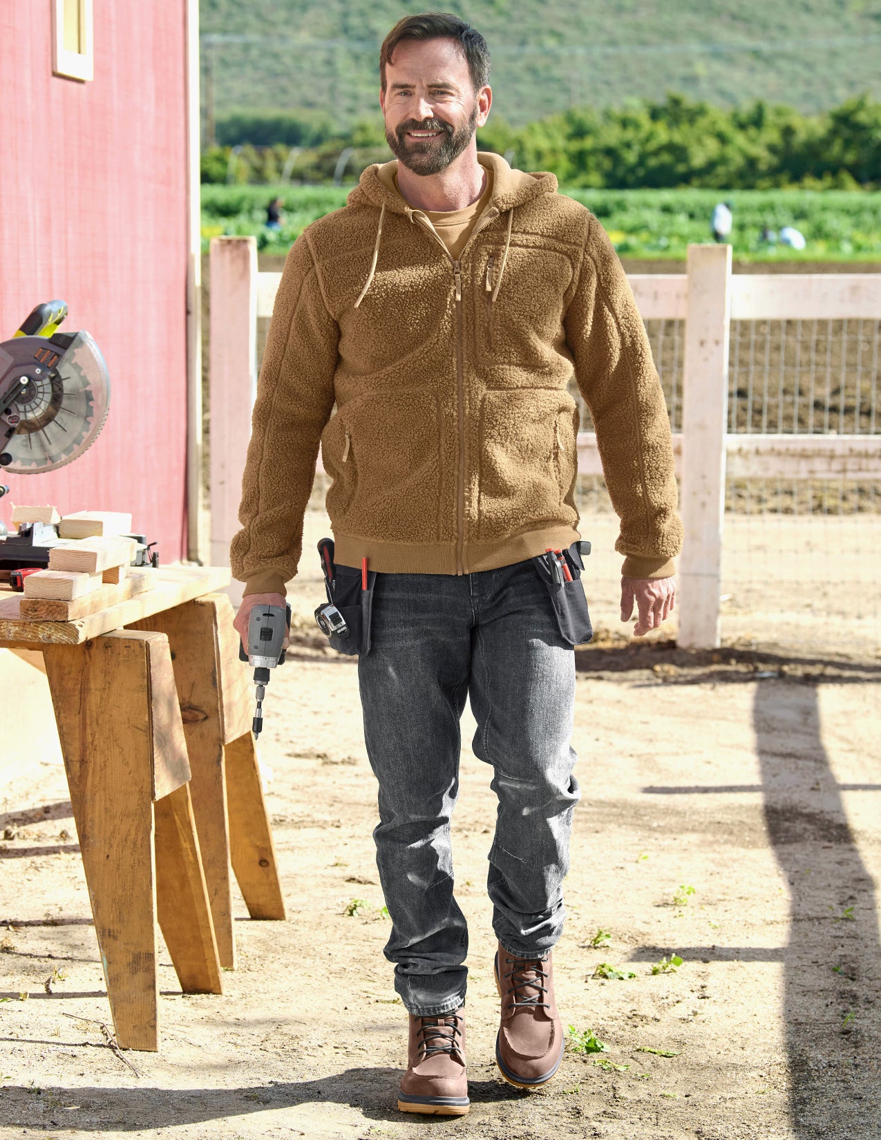 Man wearing a brown fleece hoodie and jeans standing outdoors near a wooden workbench.