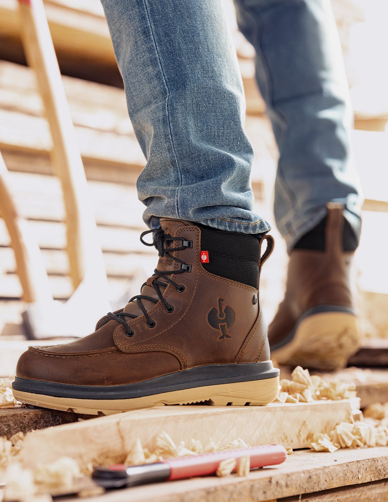 Brown work boots with a visible brand logo worn by a person on wooden planks.