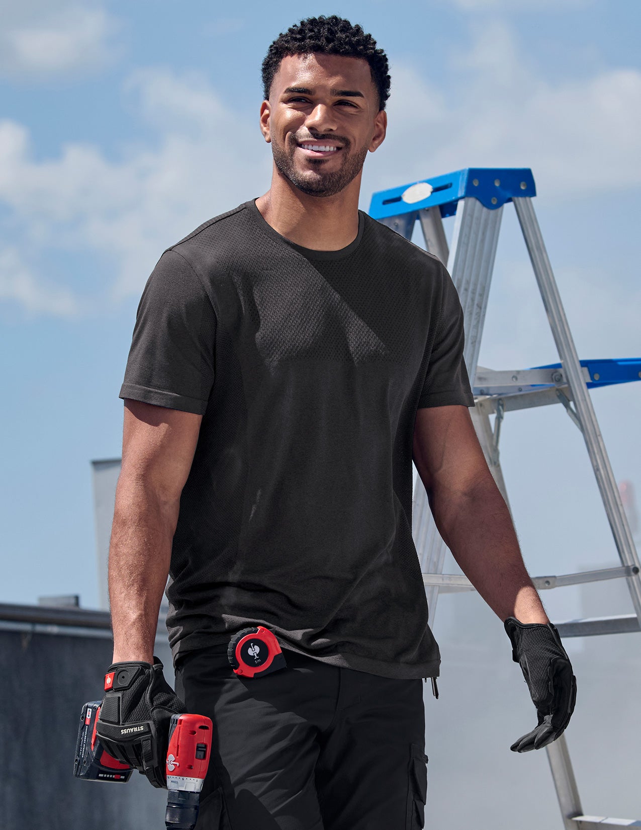 Man in black t-shirt and gloves holding a tool, with a ladder in the background.