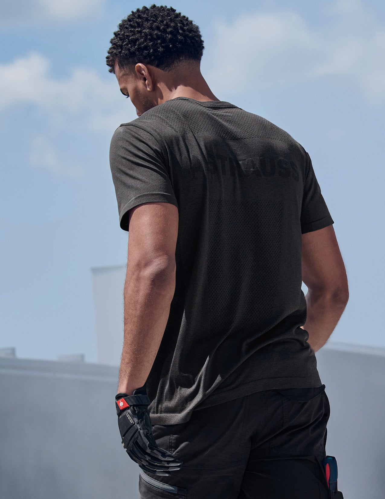 Person wearing a black t-shirt with text on the back, standing outdoors with a cloudy sky.