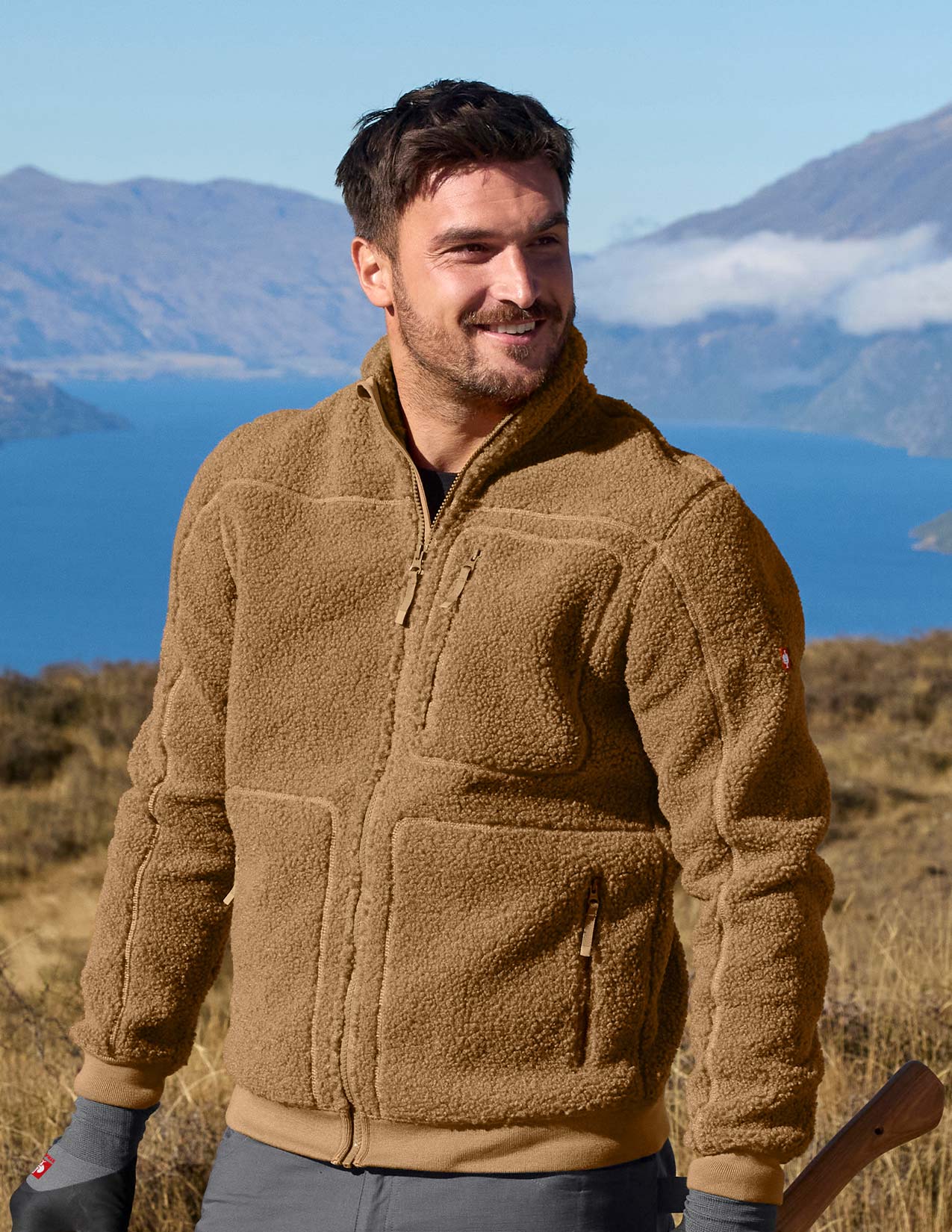Man wearing a brown fleece jacket with a scenic background of mountains and water.