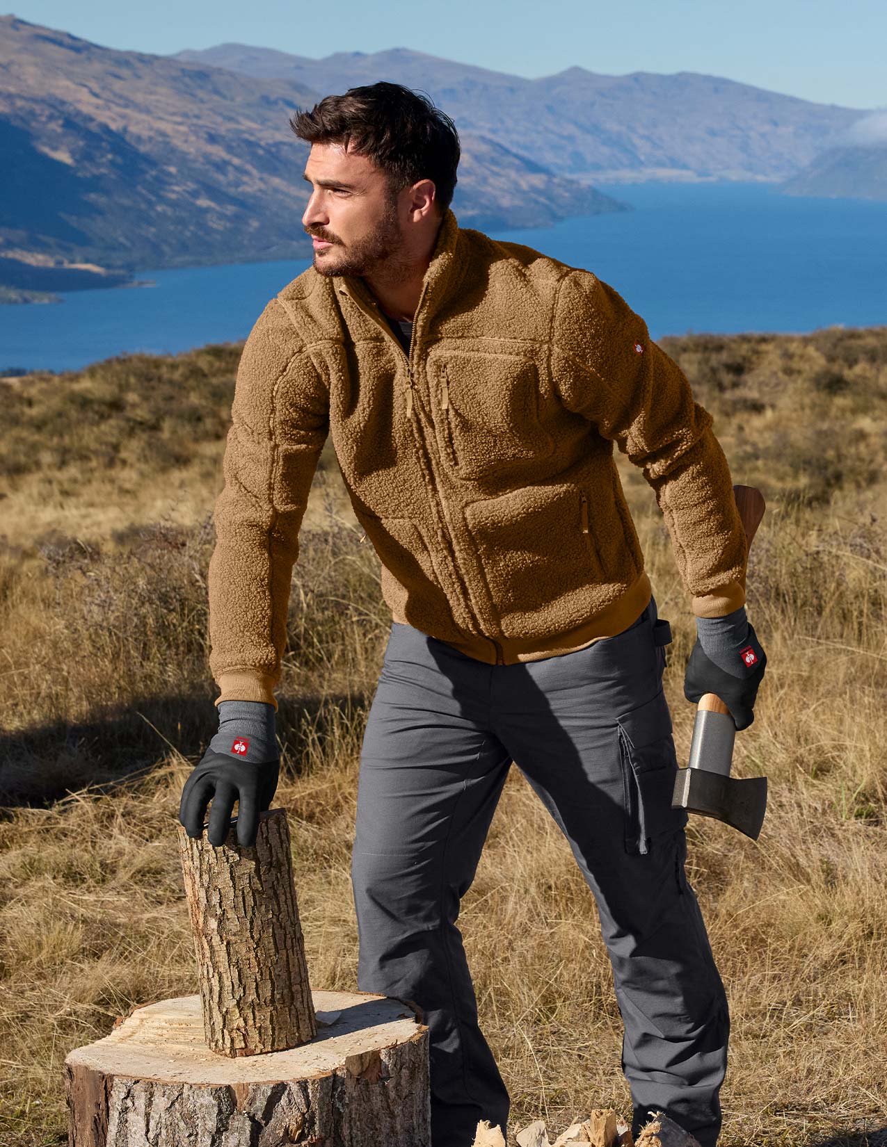 Man in a brown fleece jacket and gray pants holding an axe, standing in a scenic outdoor setting with mountains and water.