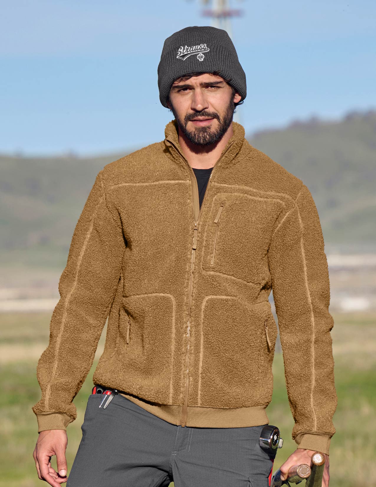 Man wearing a brown fleece jacket and grey beanie outdoors with a scenic background