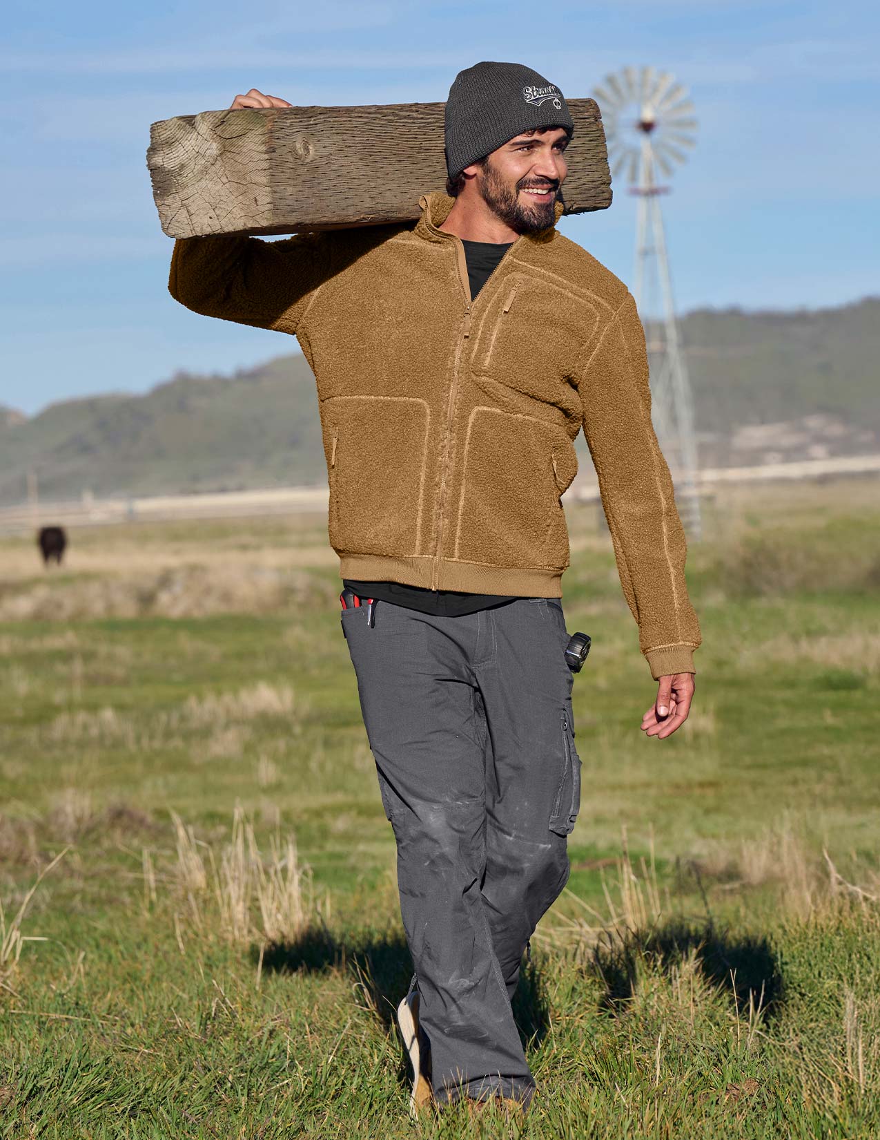 Man carrying a wooden board on his shoulder in a grassy field with a windmill in the background.